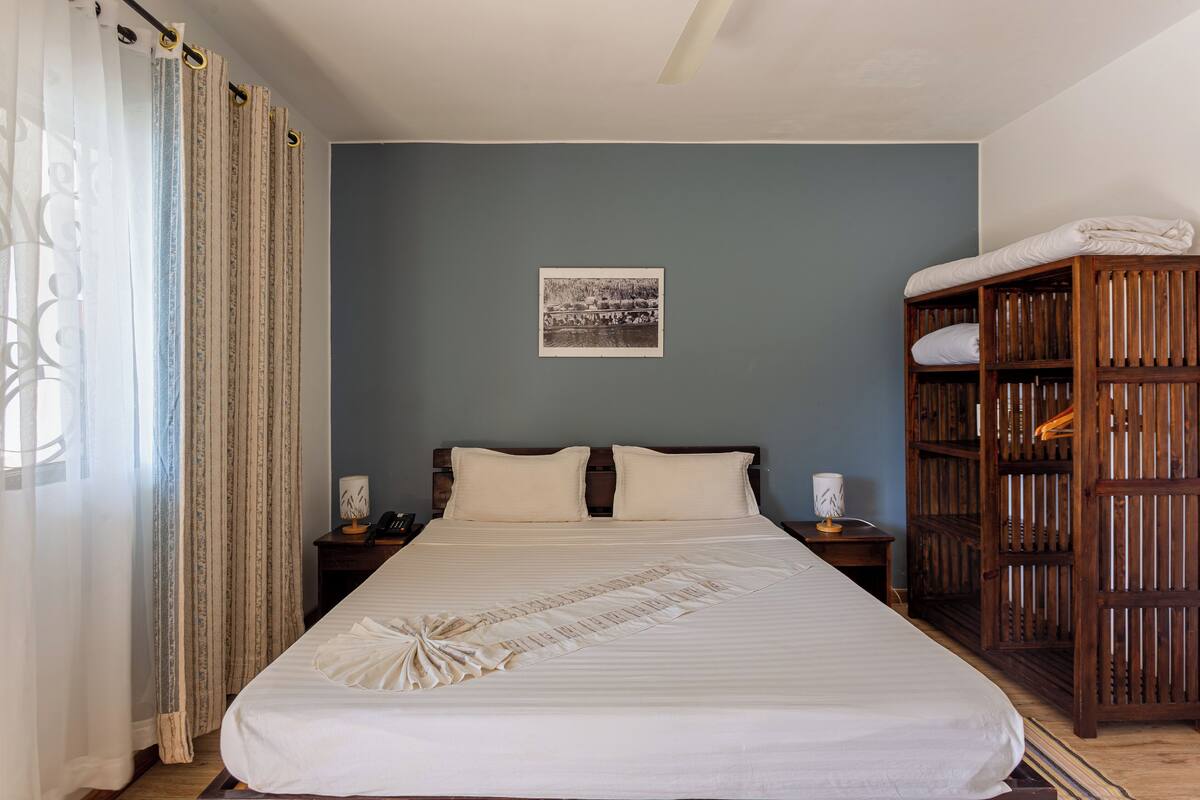 Un lit bien fait, agrémenté d’un coussin décoratif, une armoire en bois, une table de chevet avec une lampe et un tableau encadré accroché au mur.