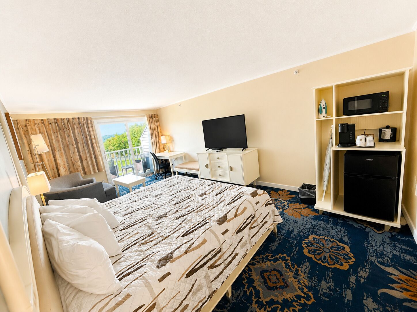 A hotel room with a bed, a desk, a TV, a minibar, and a balcony with curtains.