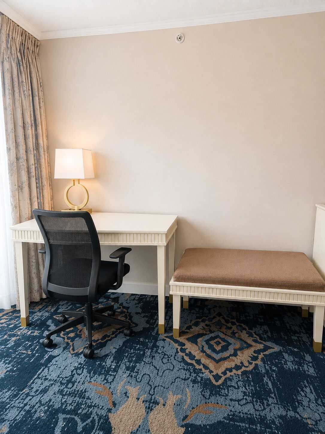 Habitación de hotel con un escritorio blanco, una silla de oficina negra, un banco marrón, alfombra estampada y una lámpara con base dorada.