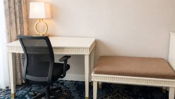 Habitación de hotel con un escritorio blanco, una silla de oficina negra, un banco marrón, alfombra estampada y una lámpara con base dorada.