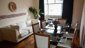 A dining area with a glass table, chairs, and a sofa. There is a window with curtains, a plant, and a wall decoration.