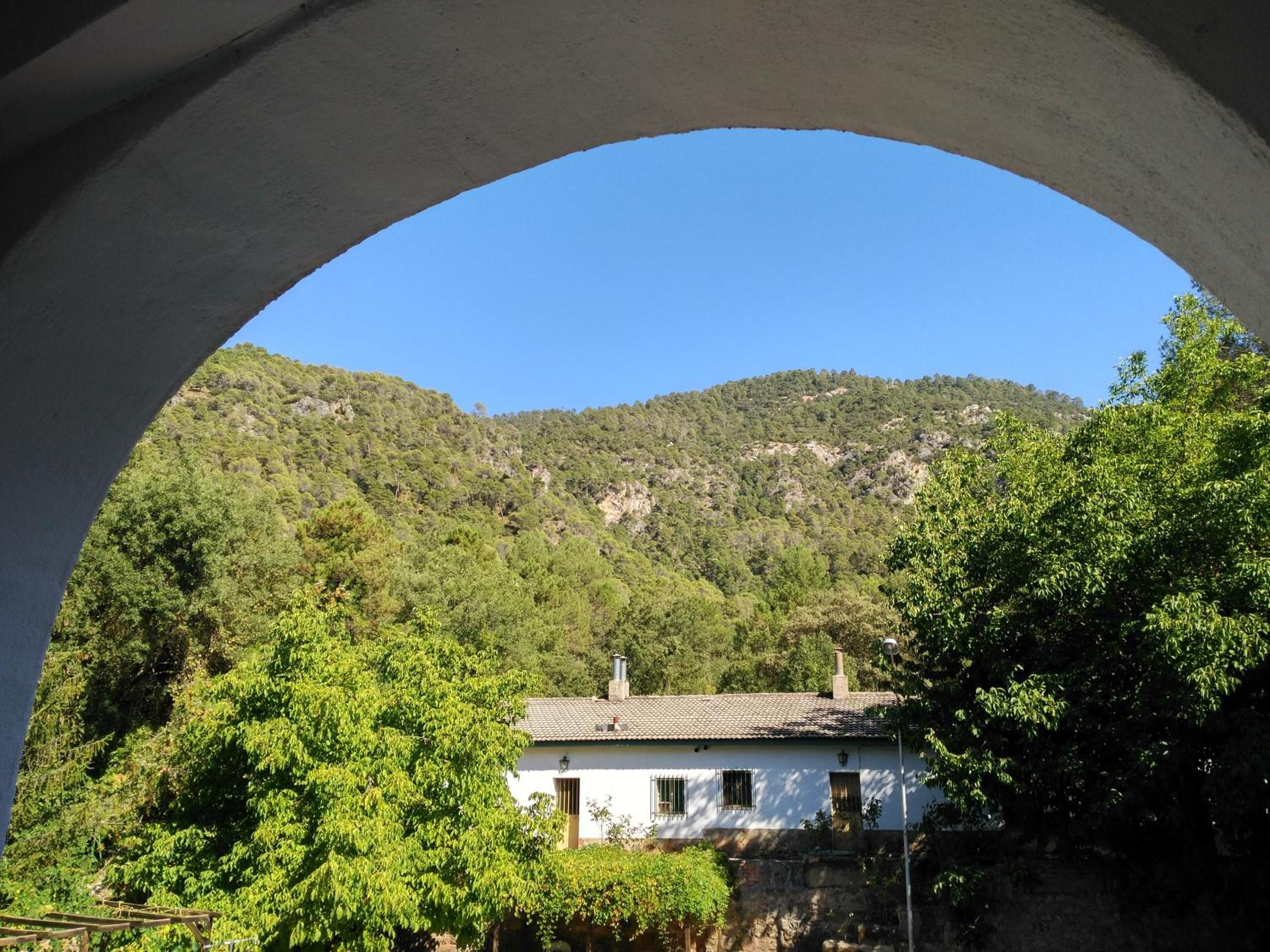 Un edificio blanco con tejado de baldosas, rodeado de abundante vegetación y un telón de fondo montañoso.