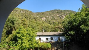 Un edificio blanco con tejado de baldosas, rodeado de abundante vegetación y un telón de fondo montañoso.
