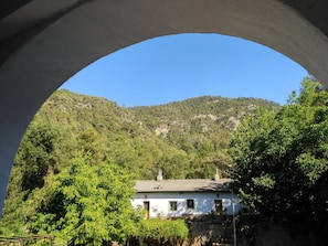Un edificio blanco con tejado de baldosas, rodeado de abundante vegetación y un telón de fondo montañoso.