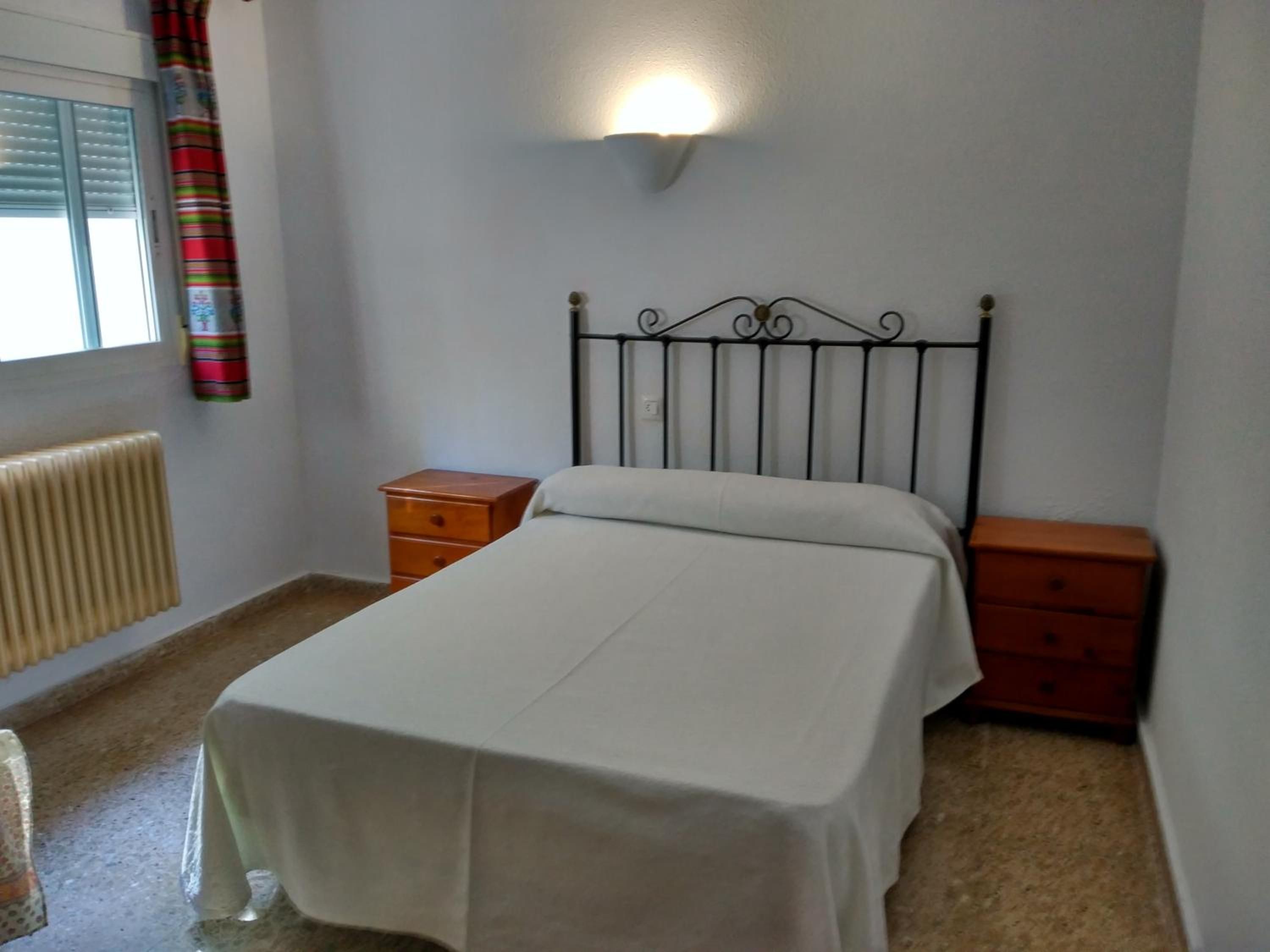 Un dormitorio con cama, mesita de noche, cómoda, radiador y ventana con cortinas.