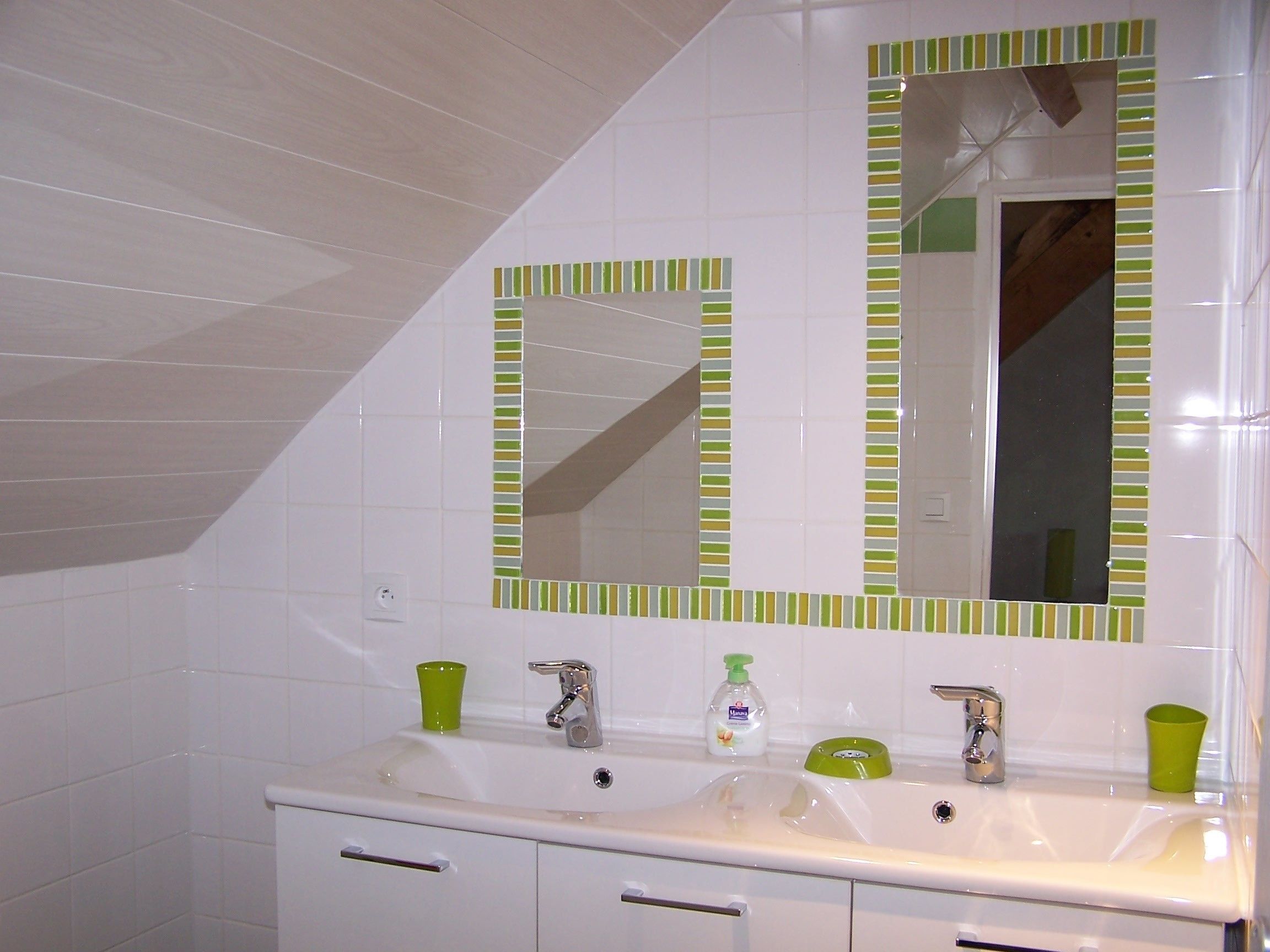 Une salle de bain avec des carreaux blancs, un lavabo blanc doté de deux robinets et un miroir à rayures vertes et blanches.