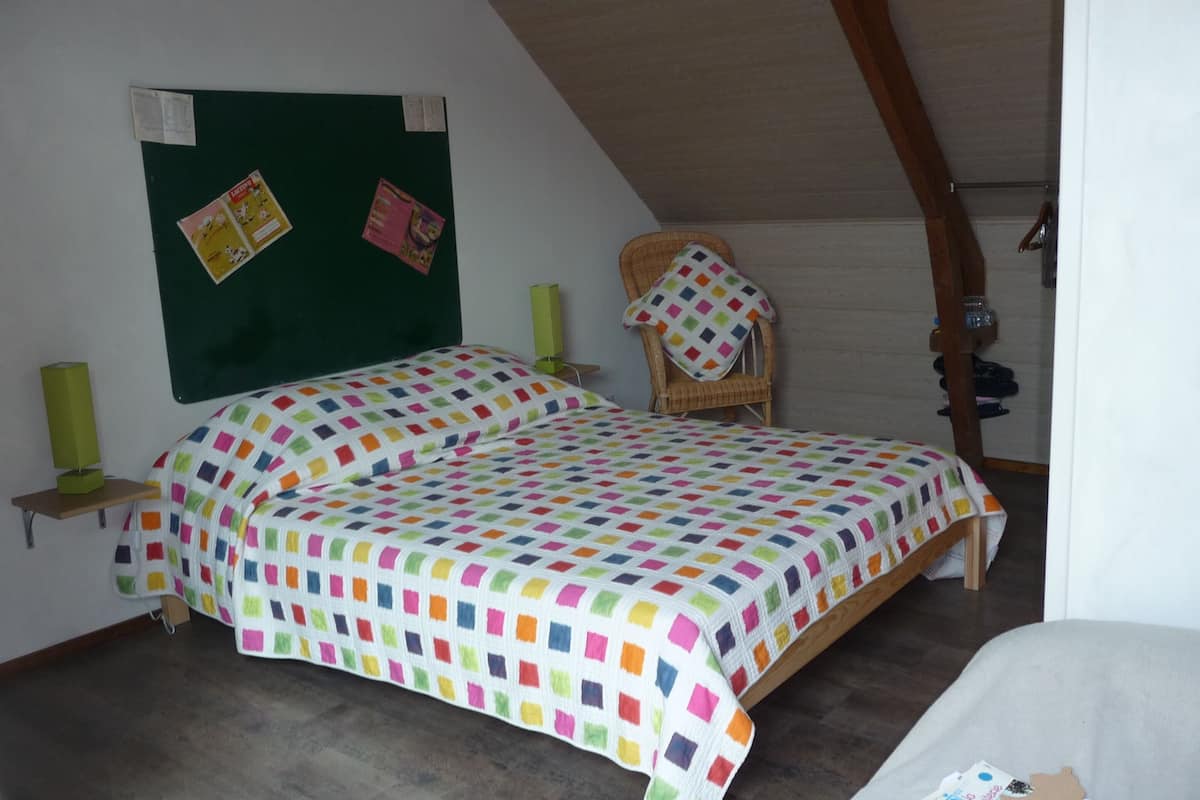 Ein Schlafzimmer mit einem Bett, einem Sessel, einer grünen Pinwand und einer Holzbalkendecke.