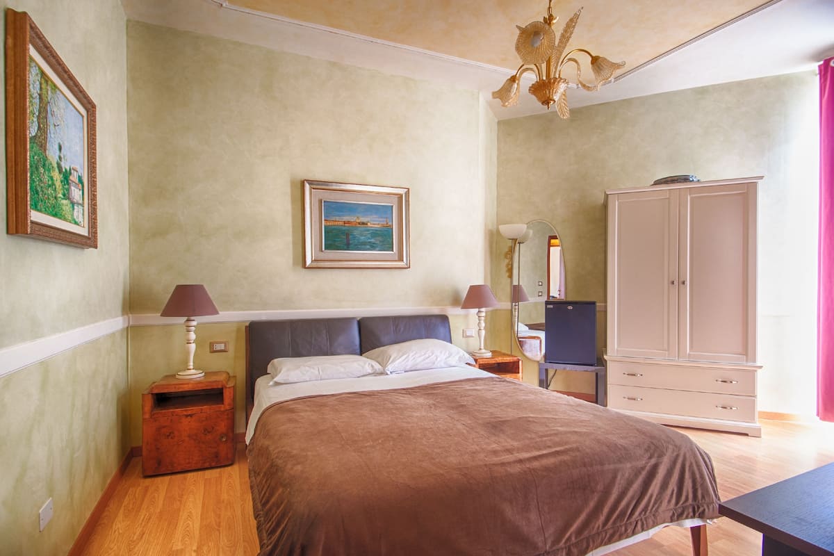 A bedroom with a bed, a wardrobe, a desk with a lamp, and a painting on the wall.
