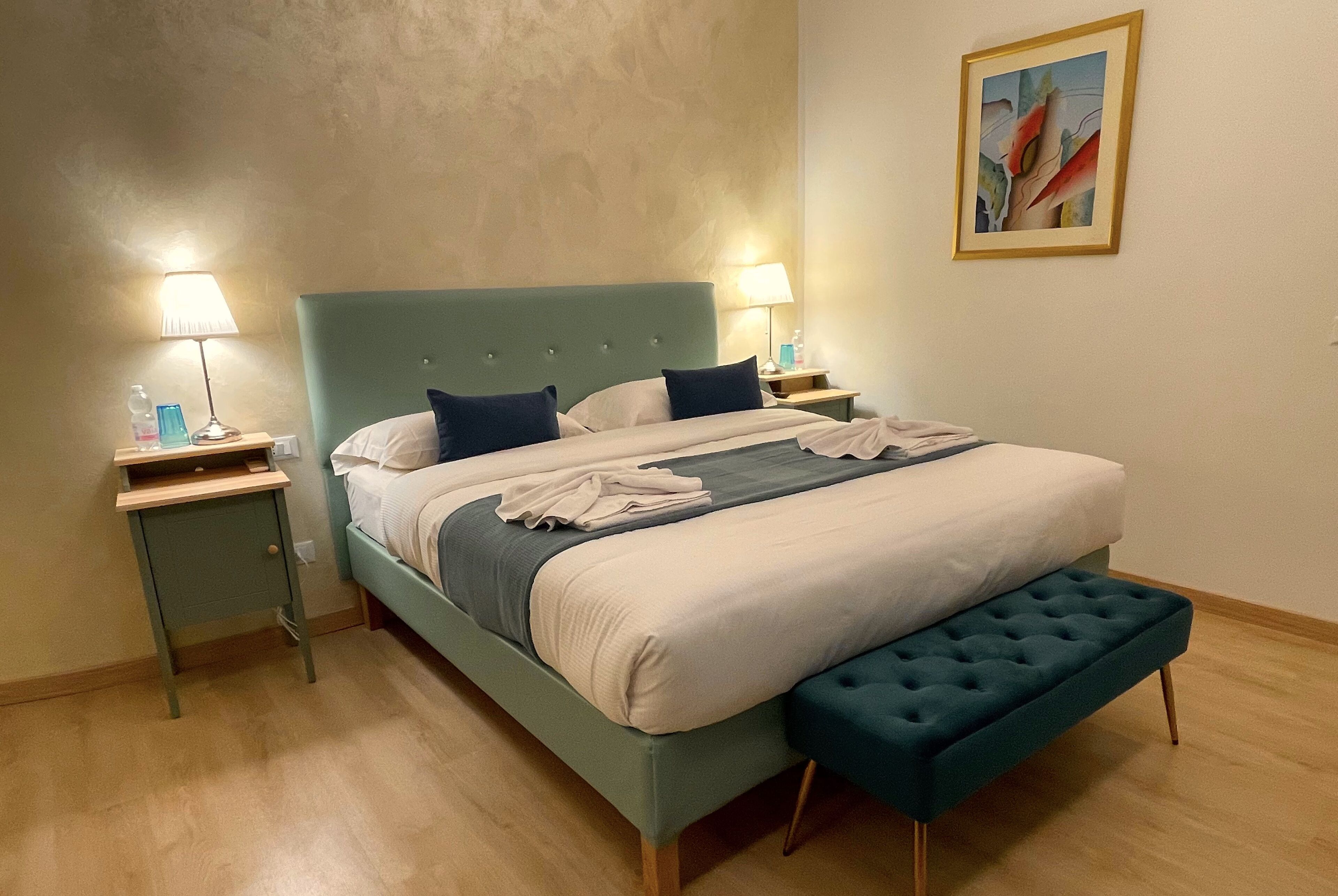 A bed with a teal headboard, two bedside tables, a bench, and a painting on the wall.
