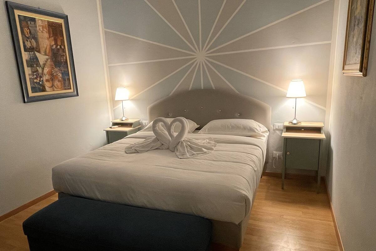 A neatly made bed with a towel heart decoration, two bedside lamps, and a framed artwork on the wall.