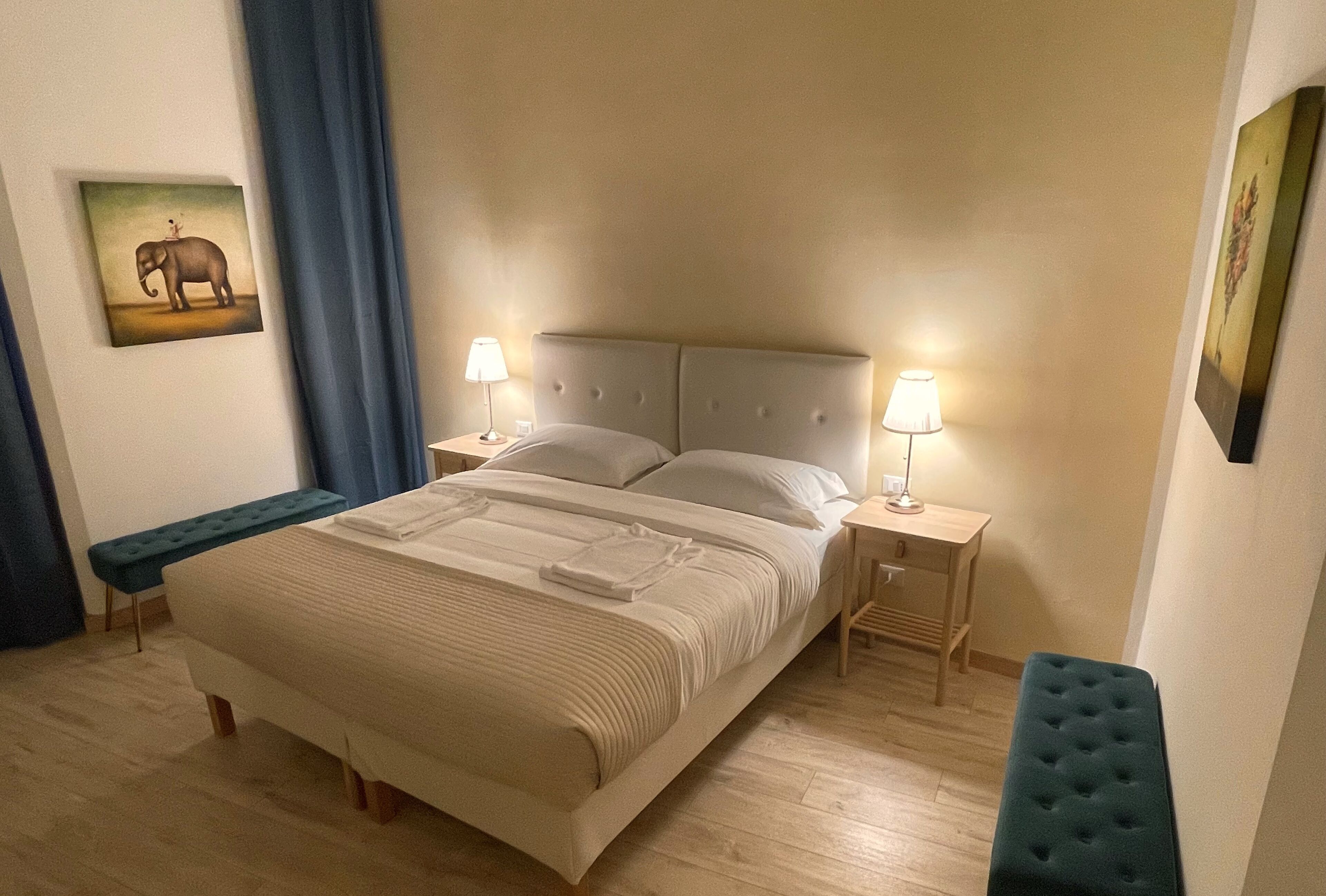 Un letto rifatto con cura, una testiera, due comodini con lampade, una panca e un quadro appeso al muro.