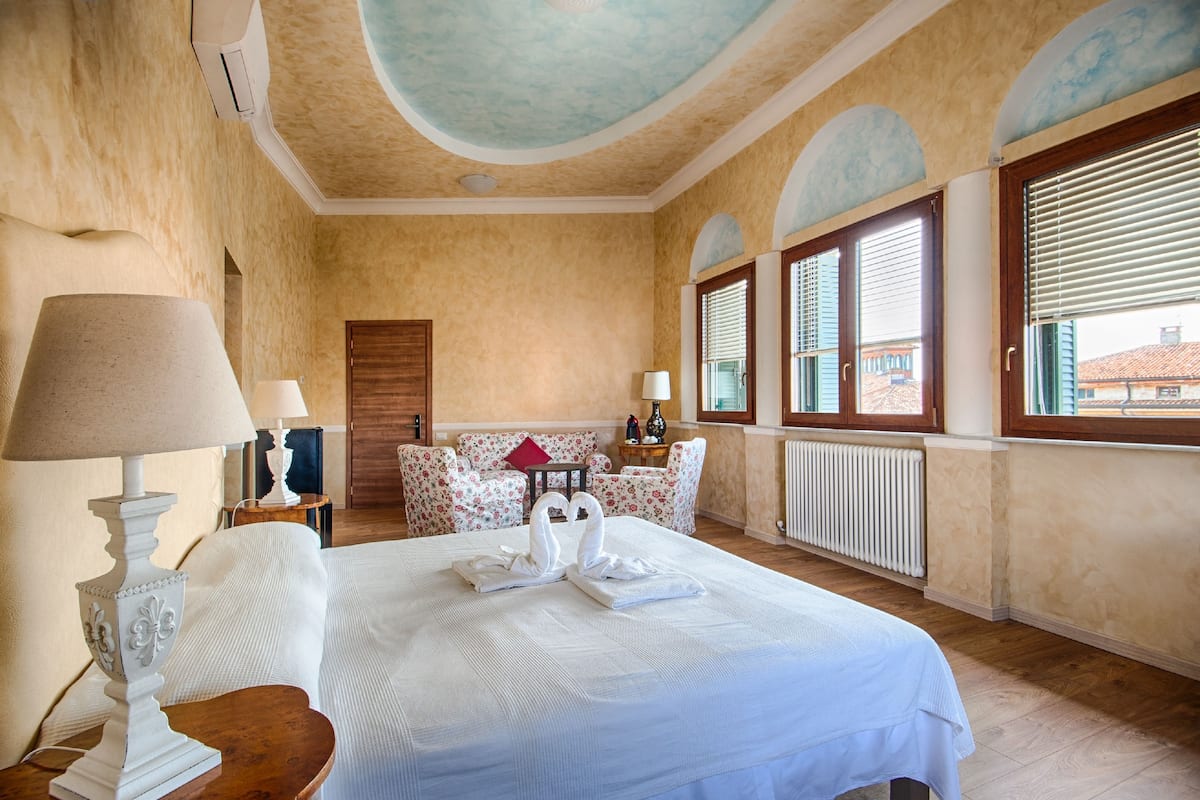 A hotel room with a bed, two lamps, a red pillow, a wooden chair, a small table, and a window with blinds.