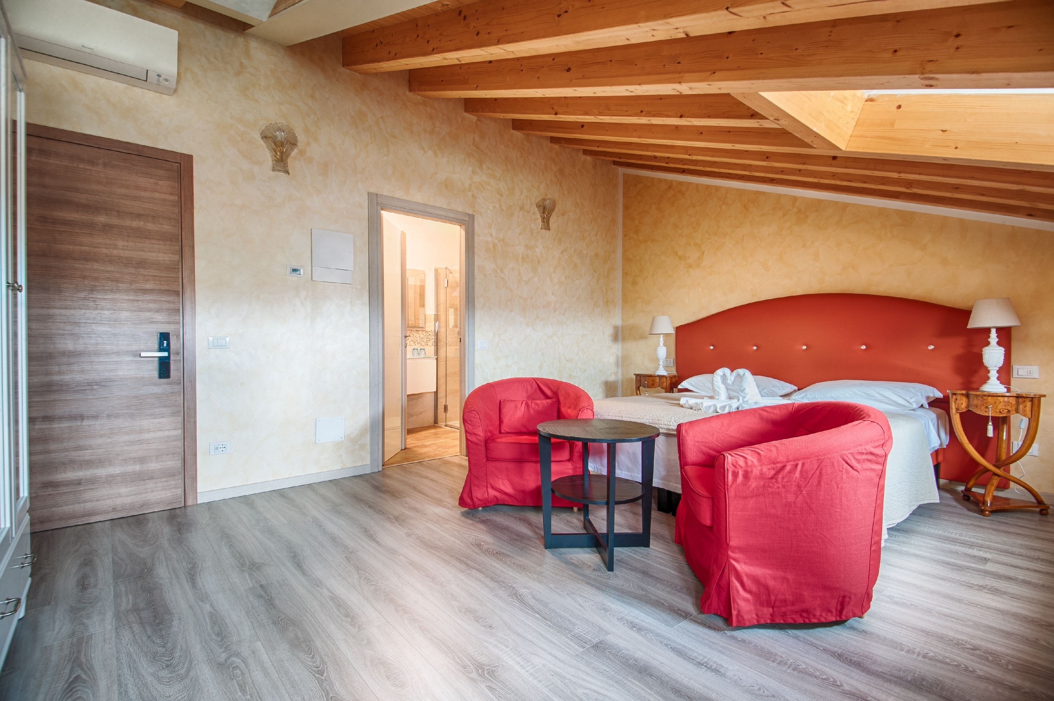Un dormitorio con techo de madera, una cama con cabecera roja, una mesita redonda pequeña y dos sillas rojas.