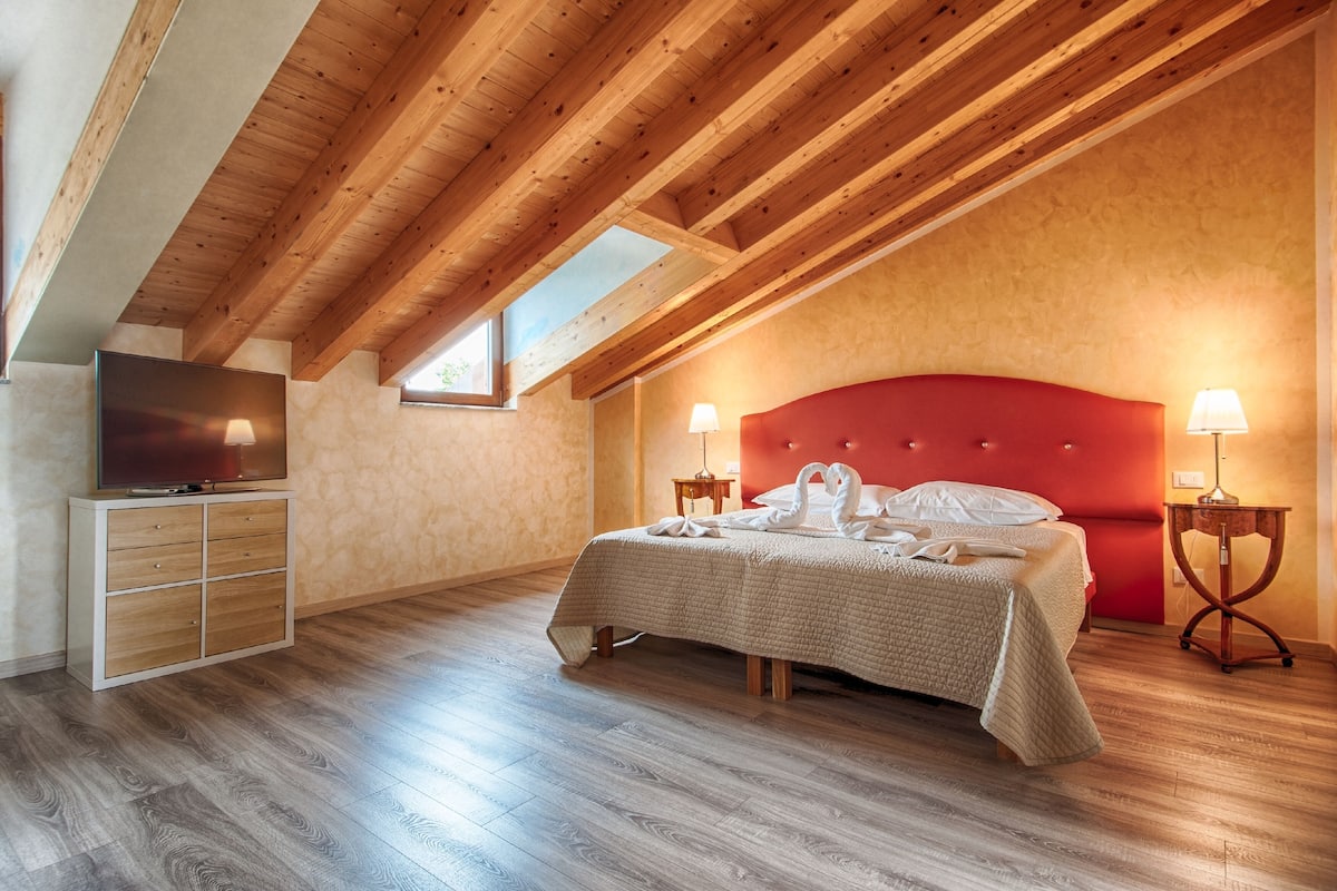 A bedroom with a wooden ceiling, a red headboard, a bed with a white blanket and two swan-shaped pillows, a TV on a wooden cabinet, and two bedside lamps.