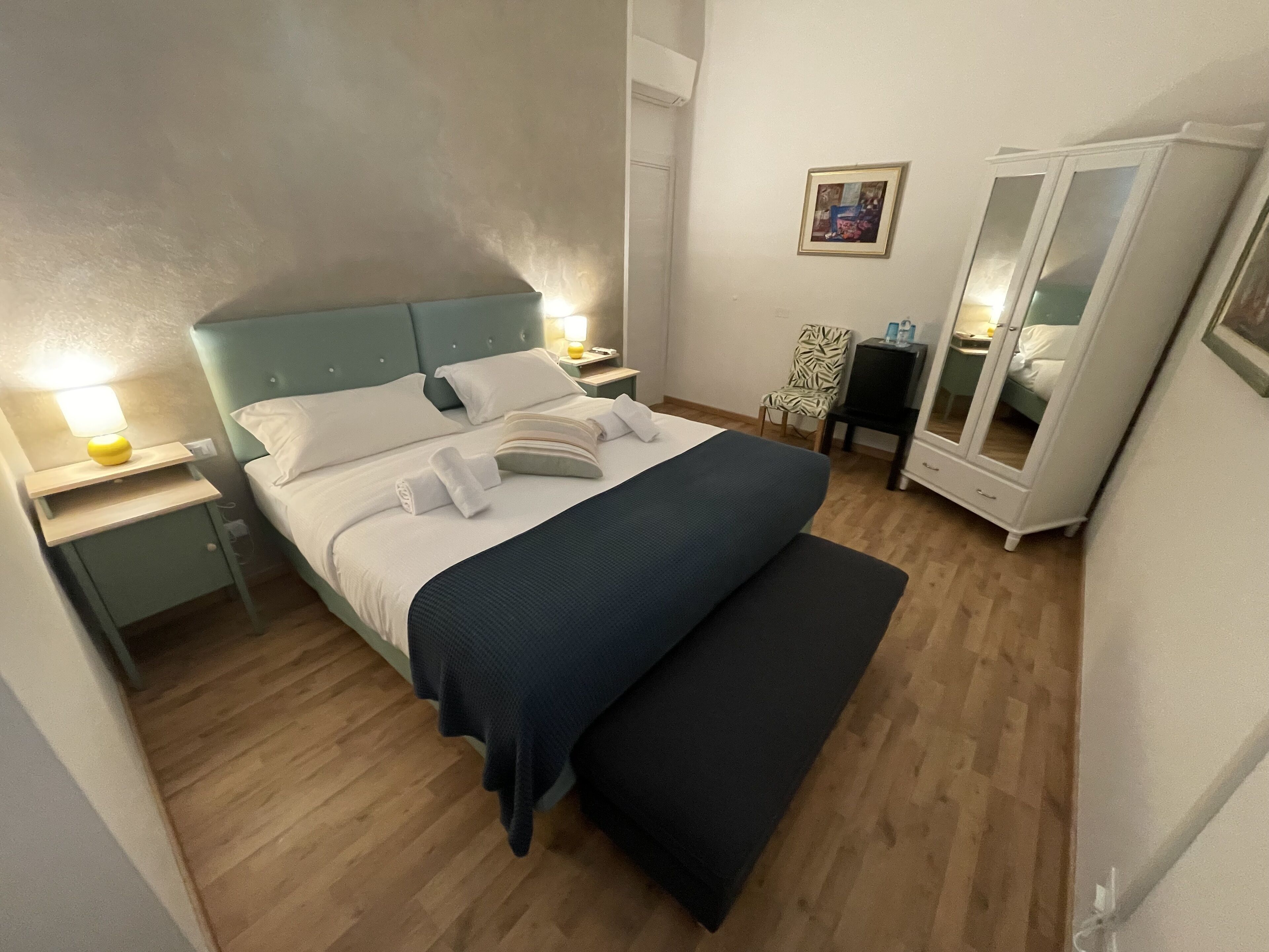 Una cama bien hecha con ropa de cama blanca y azul oscuro, una mesita de noche con lámpara, una silla, un armario y un cuadro en la pared.