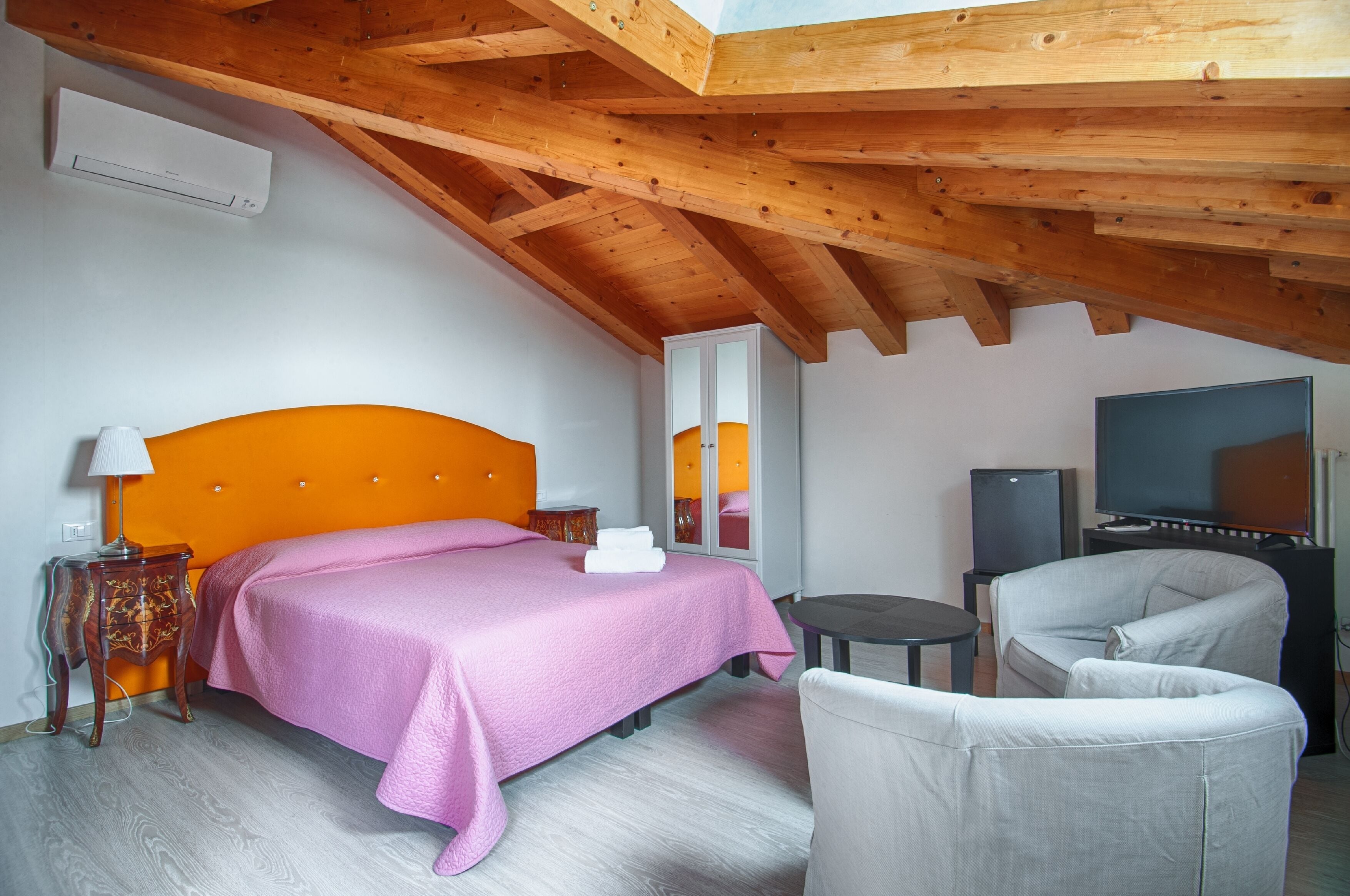 Een slaapkamer met een houten plafond, een bed met een roze dekbedovertrek, een televisie en een stoel.
