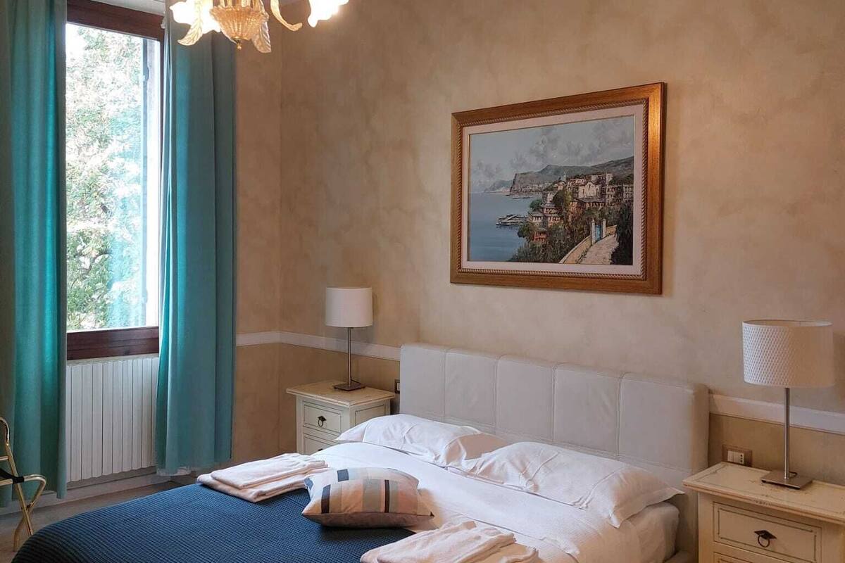 A hotel room with a bed, two bedside tables, a nightstand, a chair, a window with curtains, a chandelier, and a framed picture on the wall.