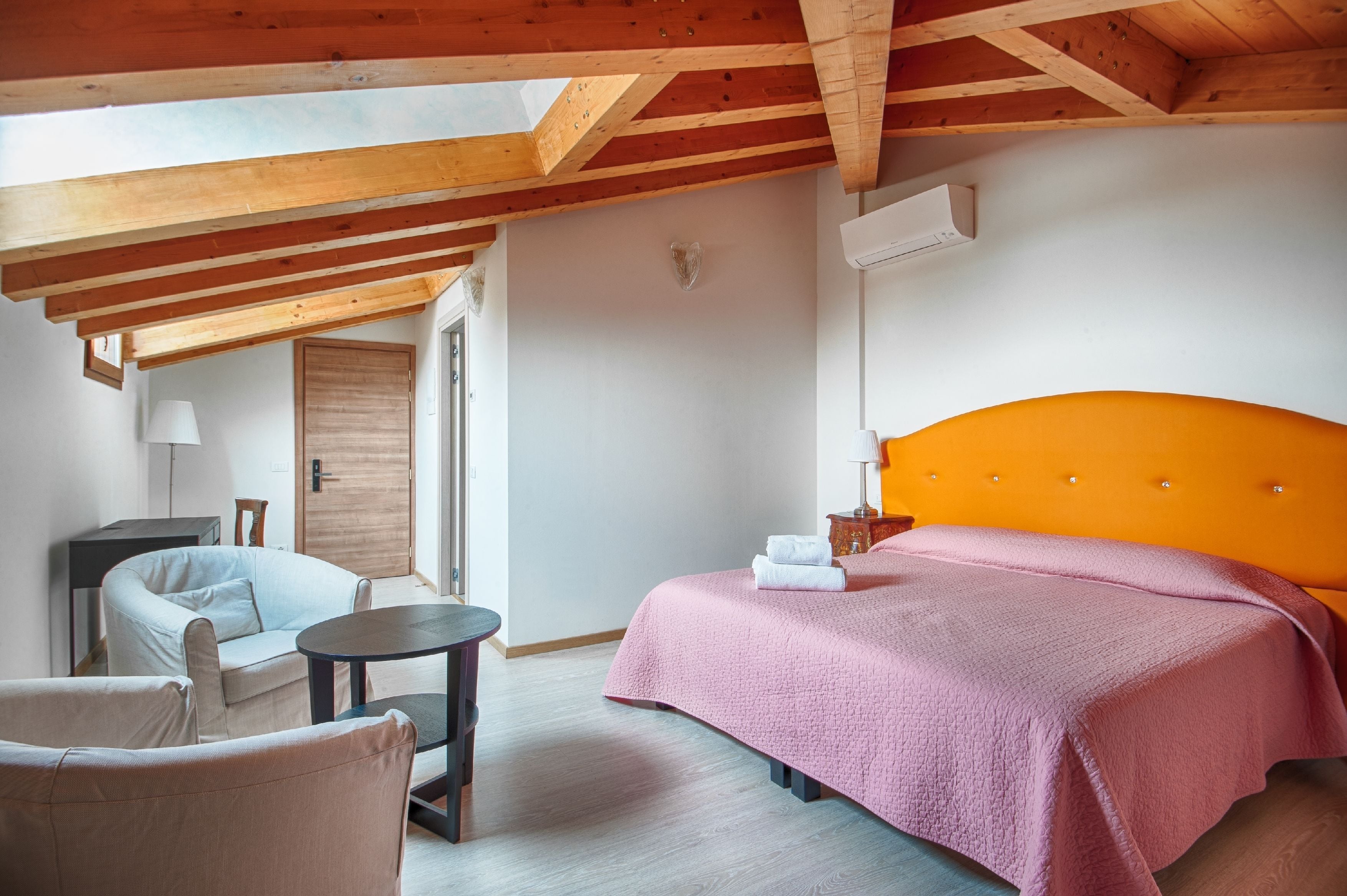 A bedroom with a wooden ceiling, a bed with an orange headboard, a pink bedspread, a small round table, and a chair.