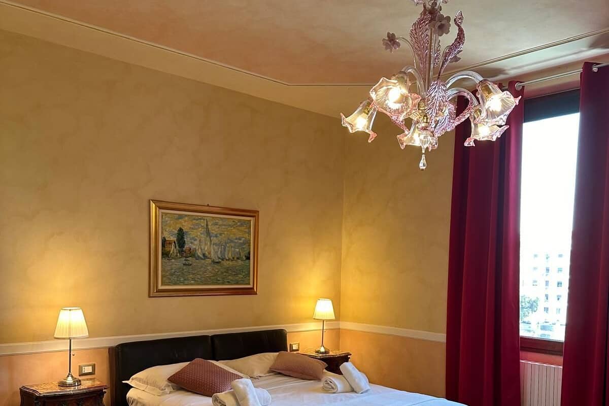 A hotel room with a large bed, two bedside lamps, a painting on the wall, a chandelier, and a window with red curtains.