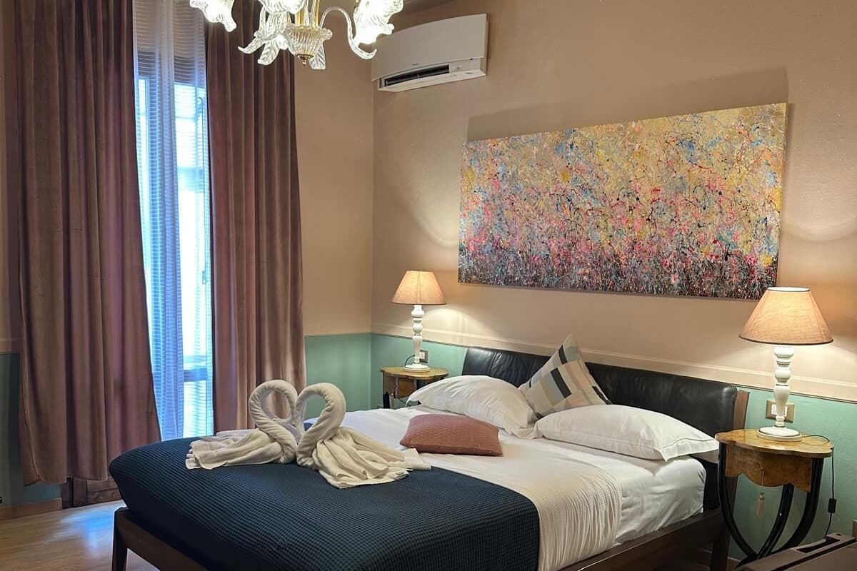 A hotel room with a large bed, two bedside tables with lamps, a chandelier, a colorful abstract painting, and a window with curtains.