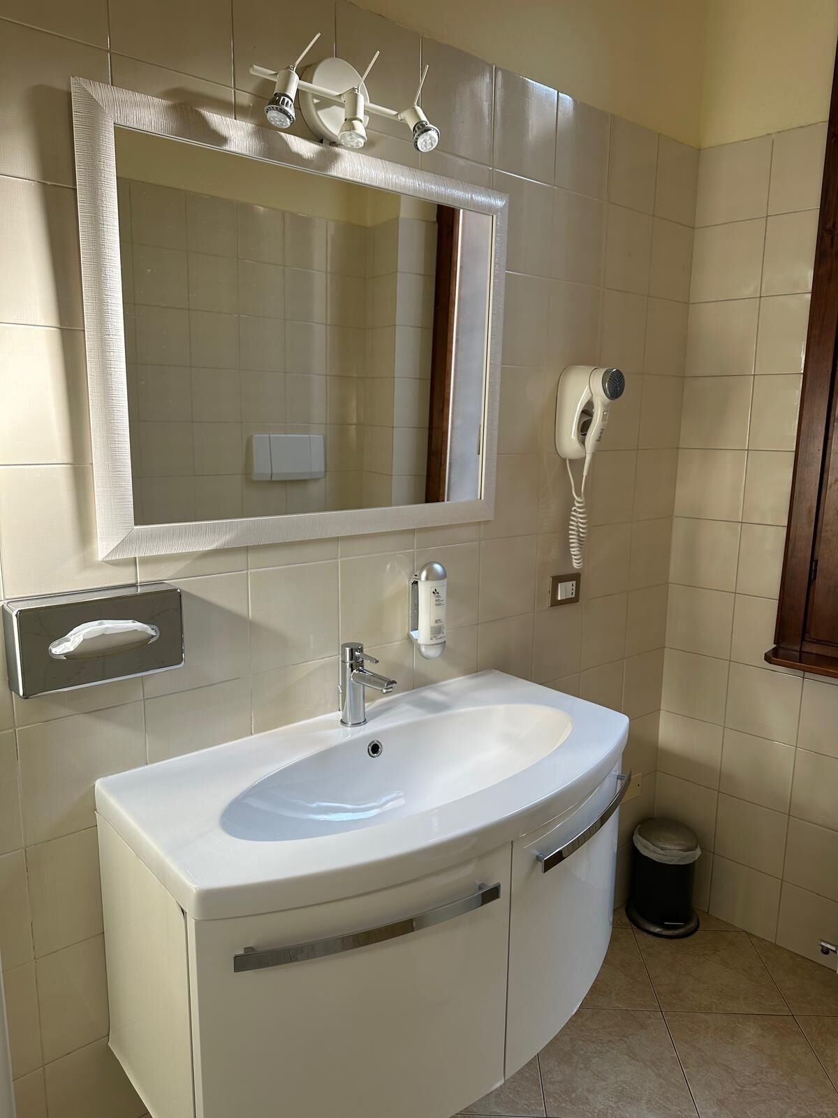 A bathroom with a white sink, a mirror with lights, a hairdryer, and a trash bin.