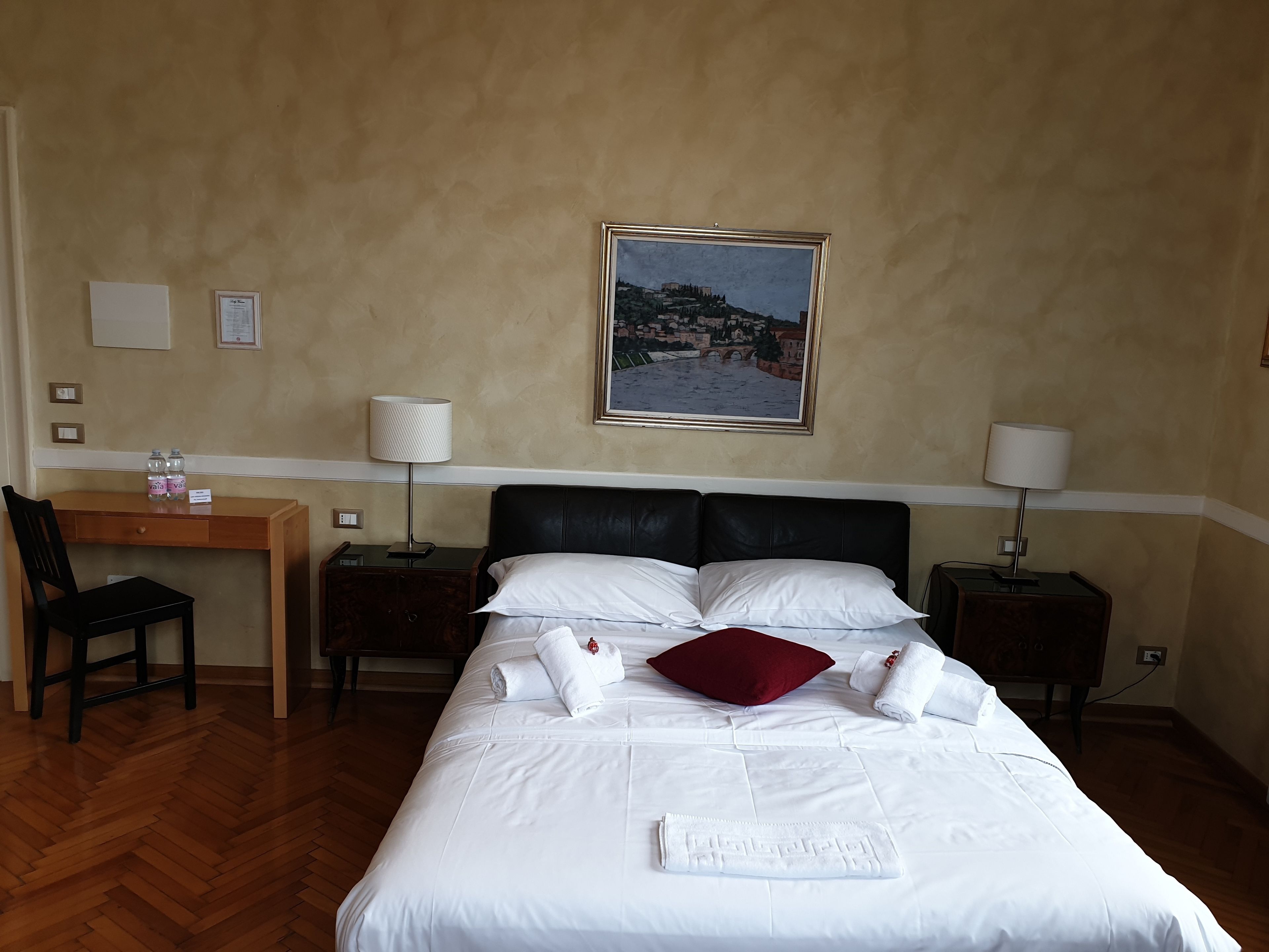 Una camera d'albergo con un letto, una scrivania, una sedia, due comodini con lampade e un quadro appeso al muro.