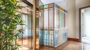 A modern elevator with a glass door, a decorative panel with a geometric pattern, and a potted plant beside it. - Lady Verona (Verona)