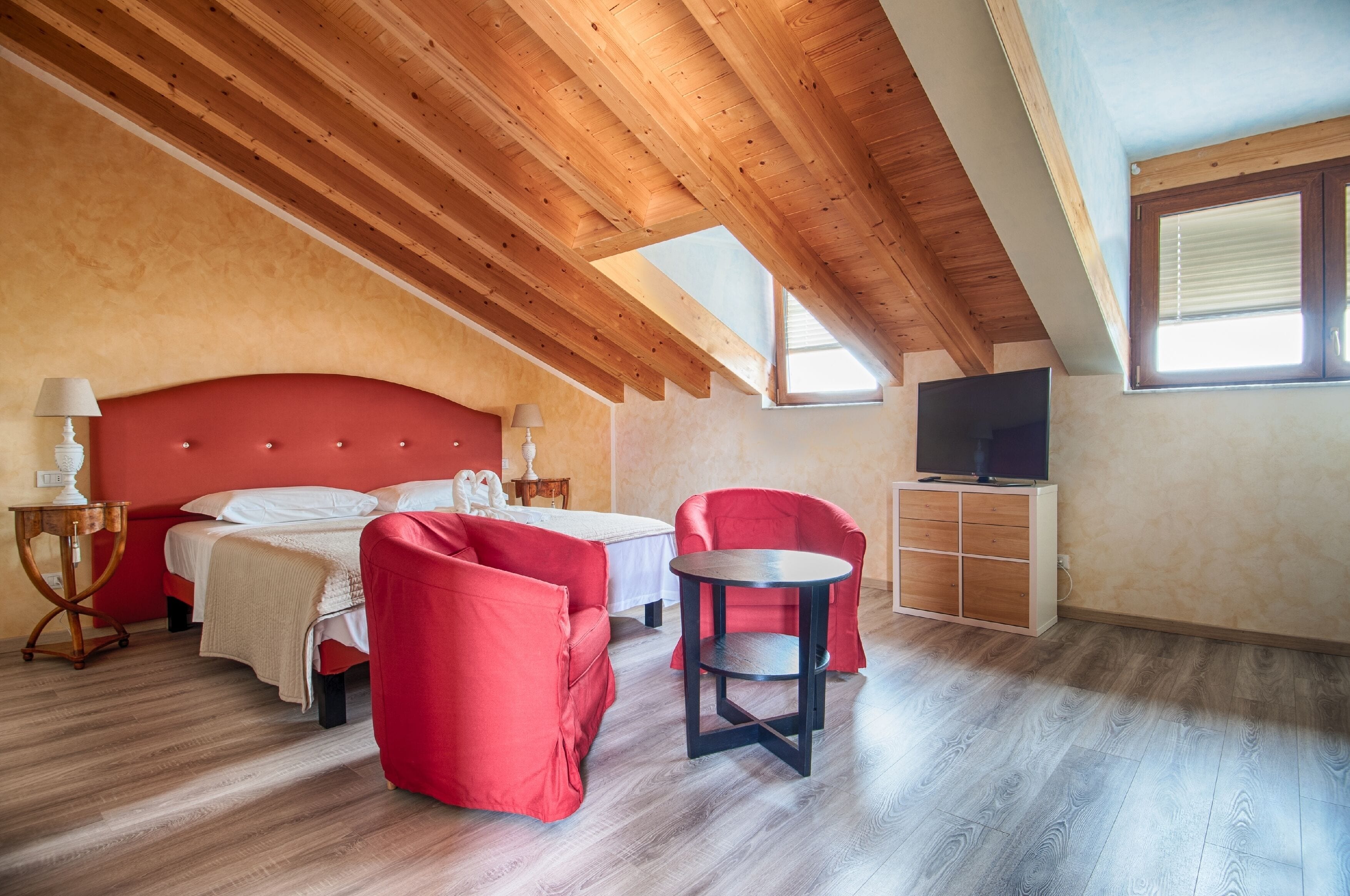 A bedroom with a wooden ceiling, a bed, a red armchair, a small table, and a TV.