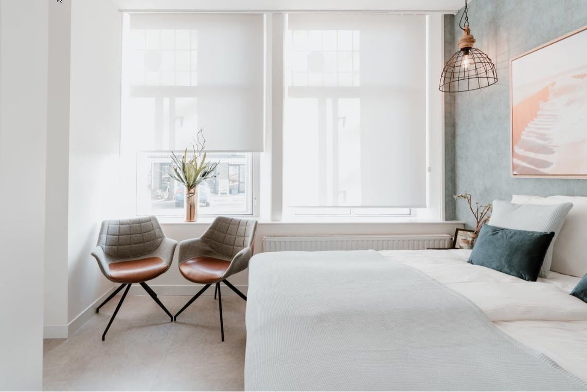 Een moderne slaapkamer met een groot bed, twee stoelen, een raam met zonwering, een radiator en een hanglamp.