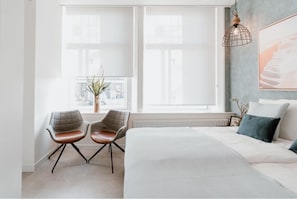 A modern bedroom with a large bed, two chairs, a window with blinds, a radiator, and a hanging light fixture. - De Zilte Melis (Meliskerke)