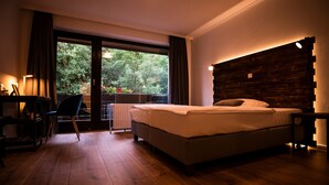 Un dormitorio con piso de madera, cama con ropa blanca, escritorio y silla, y vista a la vegetación a través de una ventana.