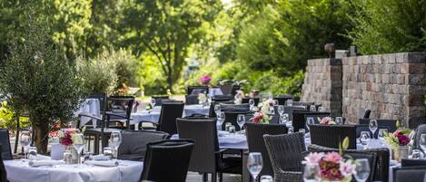 An outdoor dining area with tables set for guests, surrounded by greenery and a stone wall.
