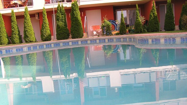A swimming pool with a tiled edge, surrounded by neatly trimmed hedges and a building with a red facade. - Gréta Wellness Apartmanok - Szt László u (Siófok)