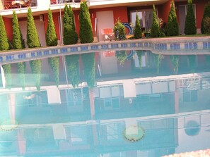 A swimming pool with a tiled edge, surrounded by neatly trimmed hedges and a building with a red facade. - Gréta Wellness Apartmanok - Szt László u (Siófok)