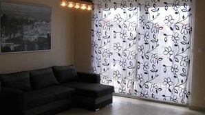 A living room with a sectional sofa, a cityscape painting, and a floral-patterned window treatment. - Gréta Wellness Apartmanok - Szt László u (Siófok)