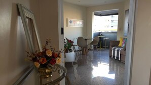 Un hall de hotel con piso pulido, una mesa consola de vidrio con un florero, un espejo enmarco, un mostrador de recepción blanco y una zona de estar con sillas y un sofá.
