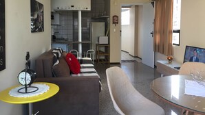 Un espacio habitable pequeño con un sofá, una mesita redonda, un televisor y una kitchenette al fondo.