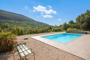 A rectangular swimming pool with a tiled deck, surrounded by gravel and plants, with a view of a hilly landscape.