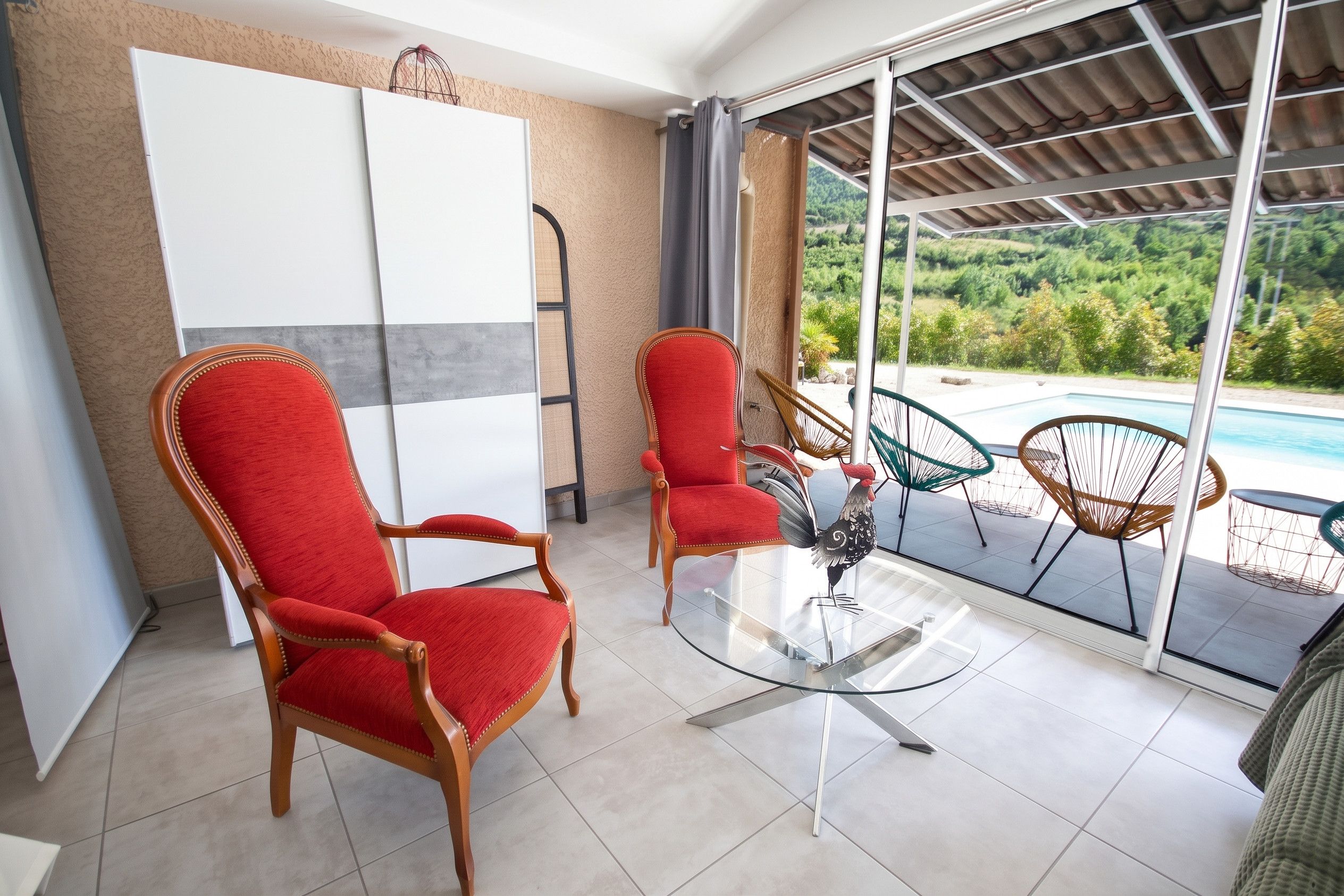 Una habitación con dos sillones tapizados en rojo, una mesa de vidrio y vistas a una piscina y vegetación al exterior.