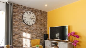 Een moderne woonkamer met een stenen muur, een groot klok, een flatscreen televisie, een bank, een bijzettafeltje en een geel poef.