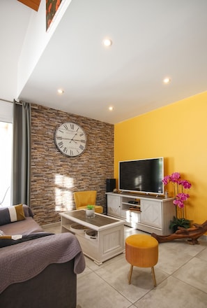 A modern living room with a stone wall, a large clock, a flat-screen TV, a sofa, a coffee table, and a yellow ottoman.