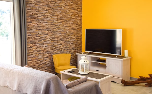 A modern room with a brick wall, a yellow accent wall, a flat-screen TV, a yellow armchair, a white coffee table, and a bed with a white duvet.