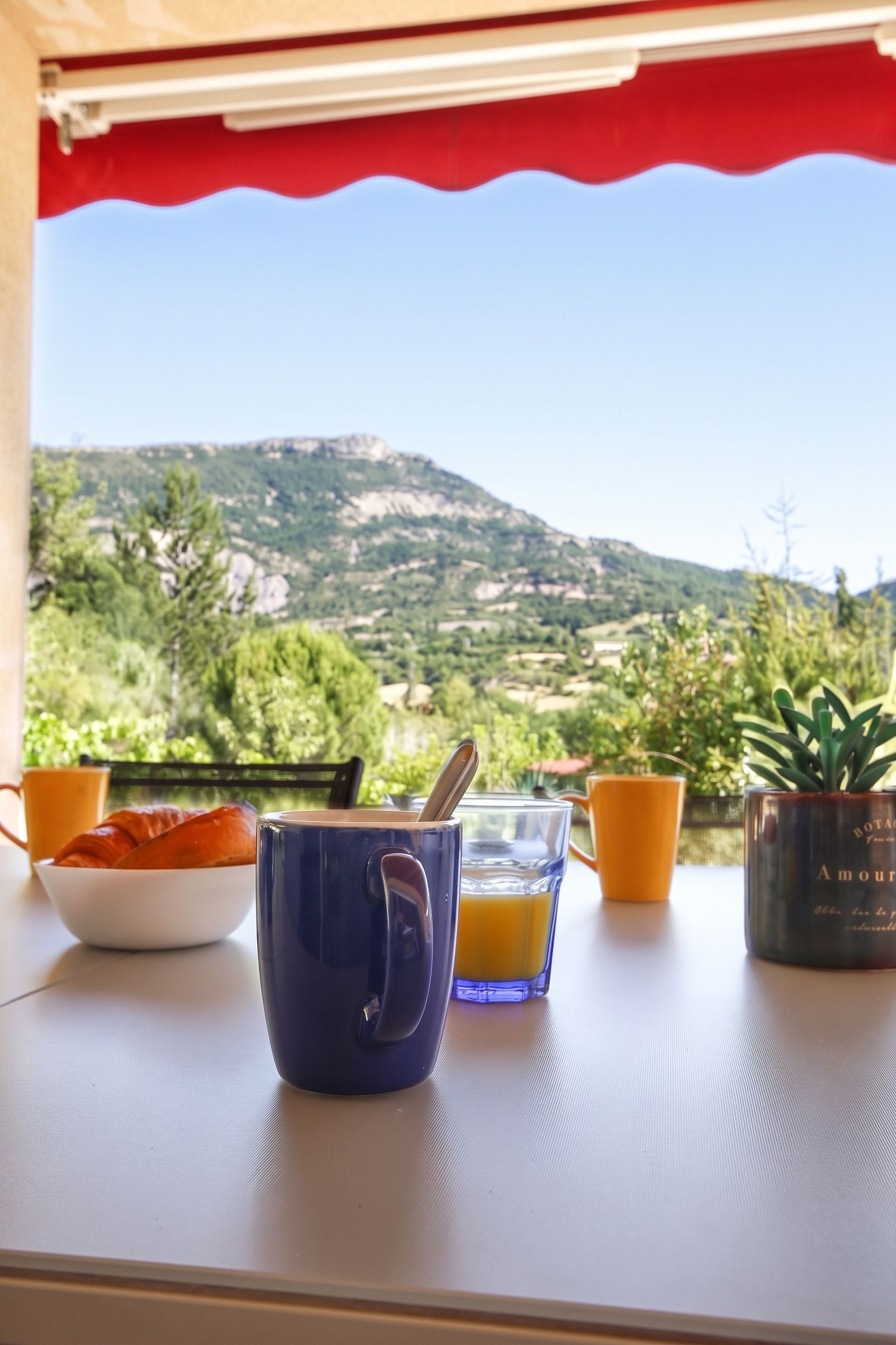 Une table avec une tasse bleue, un verre de jus d’orange et un bol de croissants, avec une vue panoramique sur les montagnes en arrière-plan.