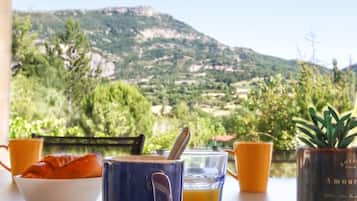 Une table avec une tasse bleue, un verre de jus d’orange et un bol de croissants, avec une vue panoramique sur les montagnes en arrière-plan.