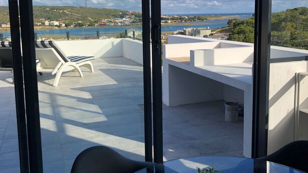 A modern rooftop terrace with white furniture, a glass table, and a potted plant. - Karee Manor Guesthouse (Still Bay)
