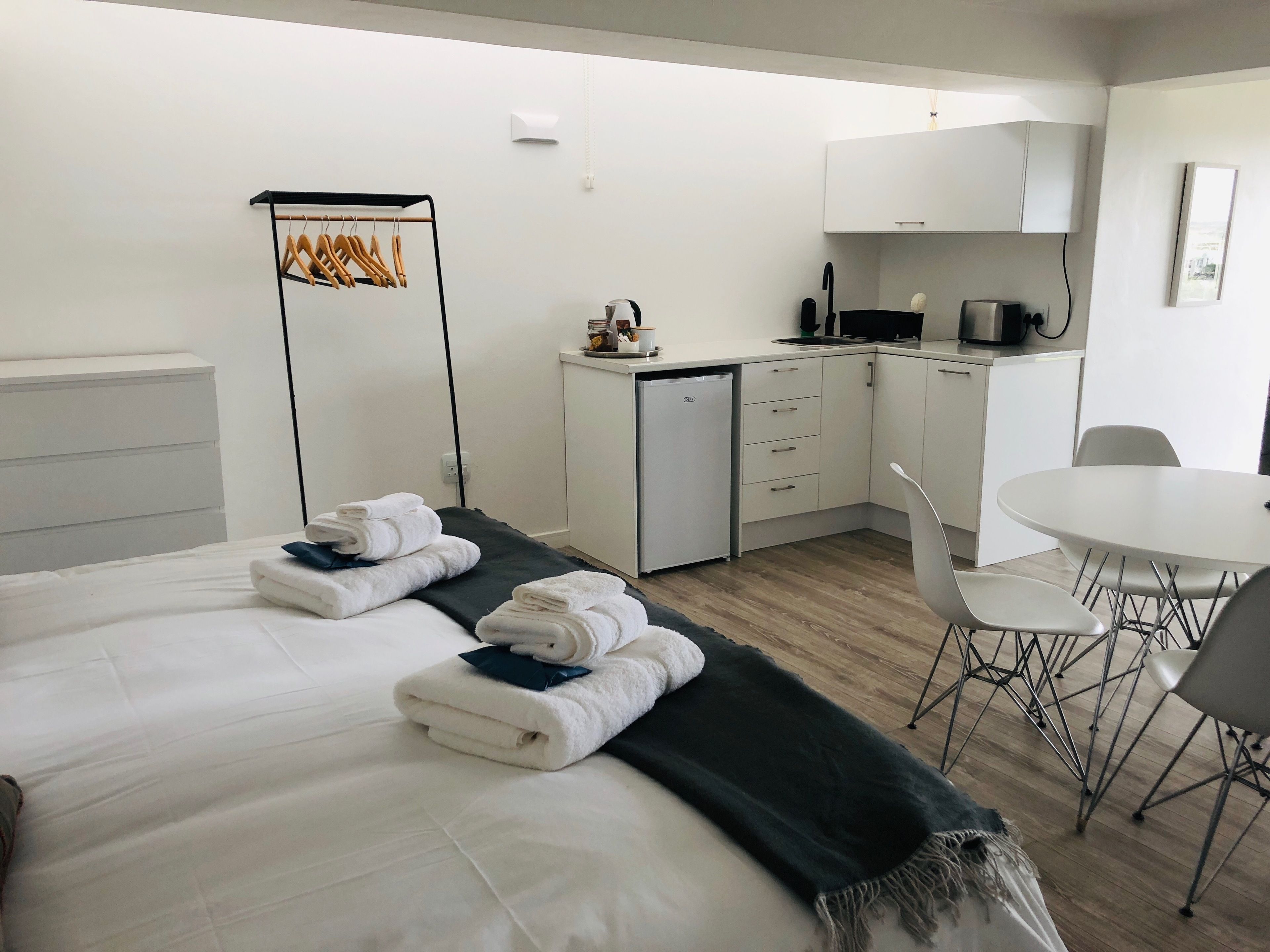 Habitación de hotel con cama, kitchenette pequeña, mesa de comedor con sillas y perchero con ganchos.