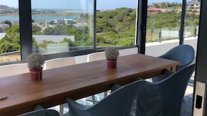 A modern room with a large window offering a view of a beach and greenery. There is a wooden table with two potted plants. - Karee Manor Guesthouse (Still Bay)