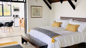 A bedroom with a large bed, a wooden bench, a desk, and a dining area with chairs. - Karee Manor Guesthouse (Still Bay)