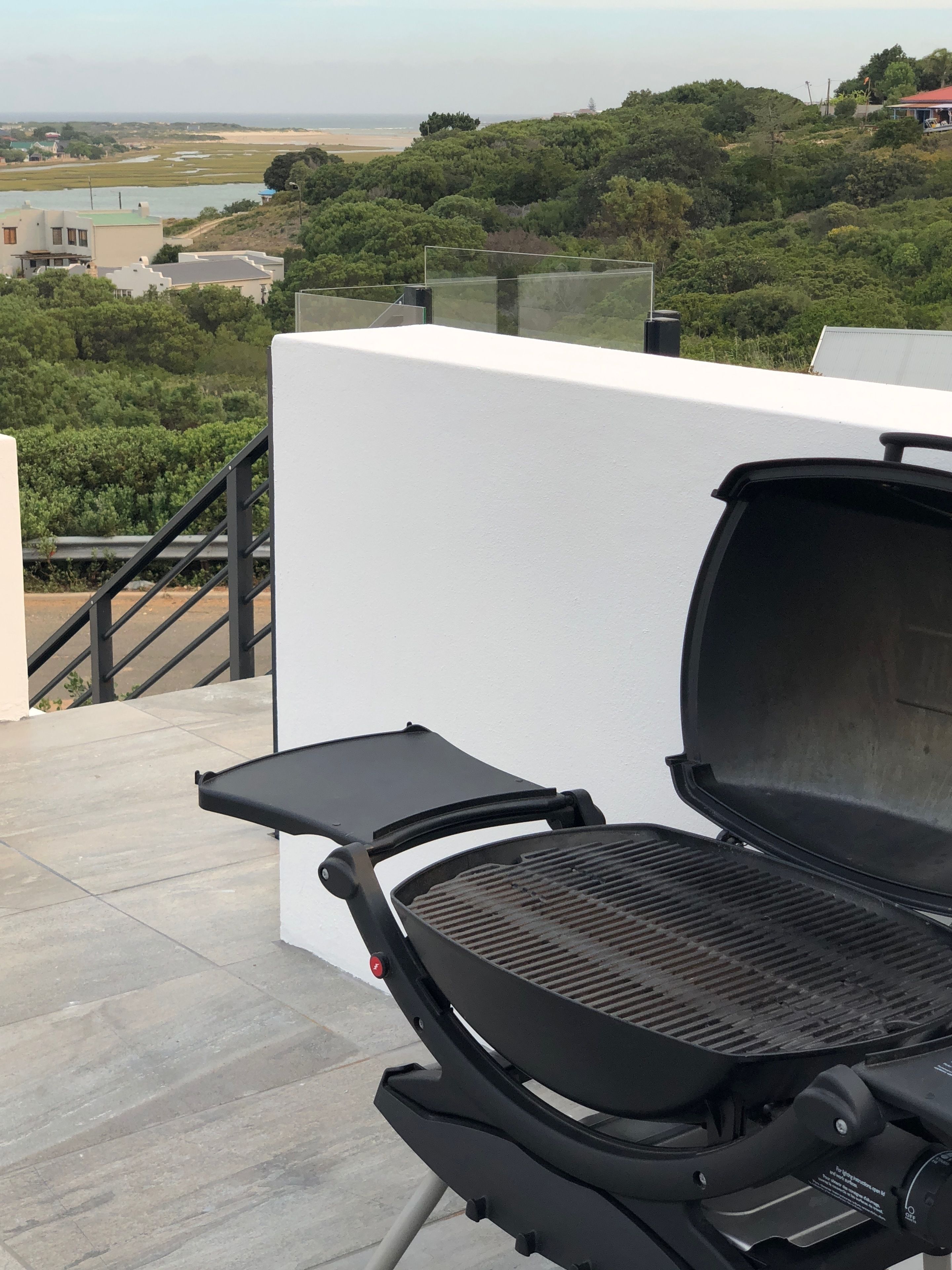 A rooftop patio with a grill, a white wall, and a view of the beach and greenery.