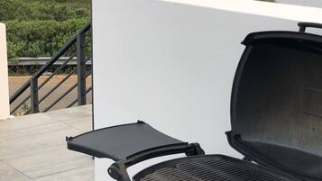 A rooftop patio with a grill, a white wall, and a view of the beach and greenery.